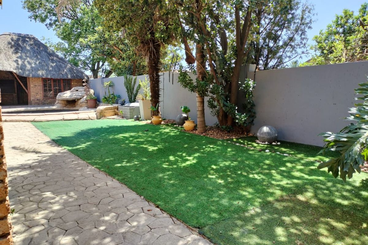 A well-maintained rear garden featuring artificial turf, paved walkways, mature trees, and a thatched-roof outdoor structure in the background.