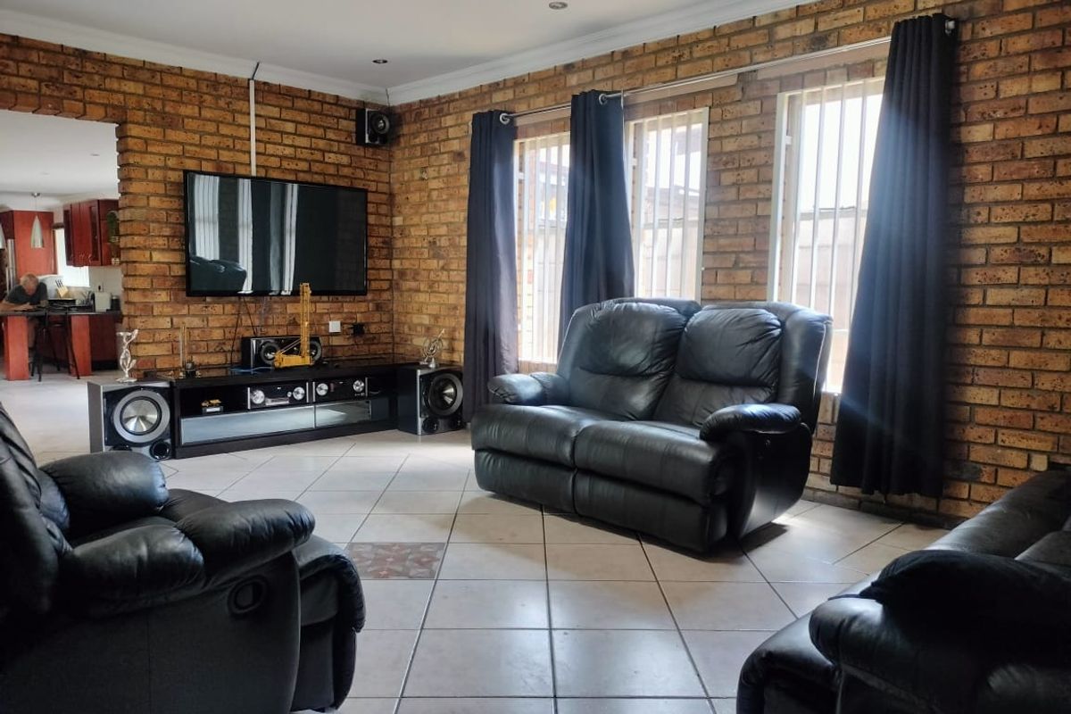 Spacious living room featuring exposed face-brick walls, tiled flooring, and large windows with security bars, leading into an adjacent kitchen area.