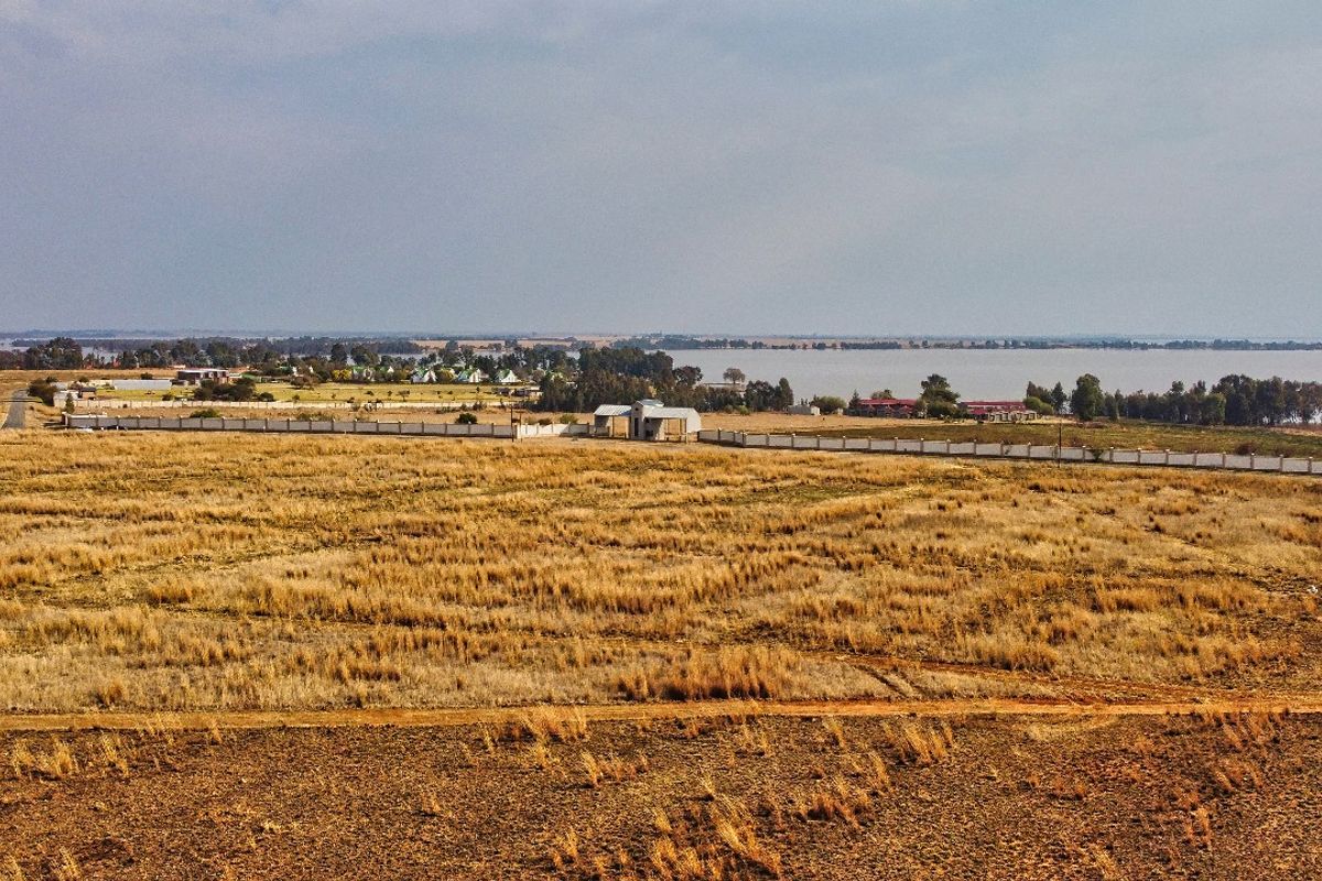 An aerial view of a large, vacant residential plot featuring perimeter fencing and a gated entrance, overlooking a nearby lake and surrounding landscape.