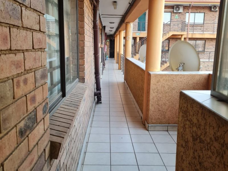 A long, covered apartment balcony featuring tiled flooring, brick wall finishes, and structural support columns, providing access to the building exterior.