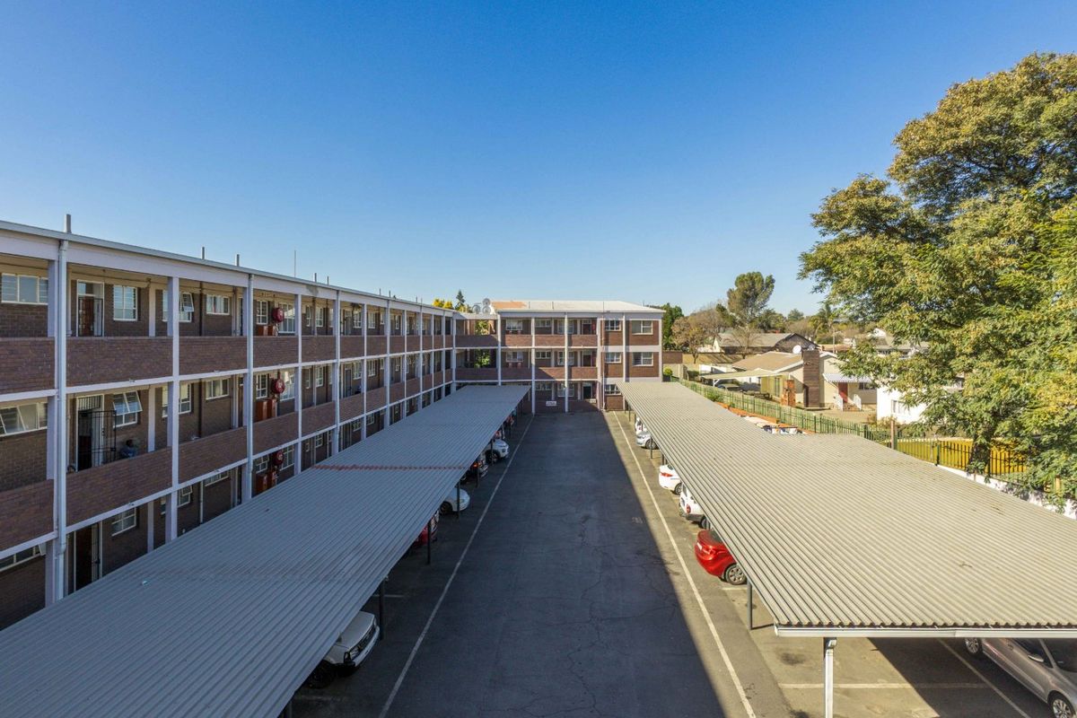 Aerial view of an apartment complex featuring a central parking area with covered carports and multi-story brick residential blocks with balconies.