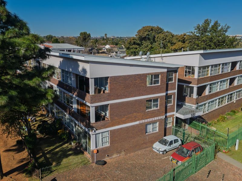 An aerial view of a multi-story brick apartment building featuring a paved driveway, secure perimeter fencing, and established surrounding trees.