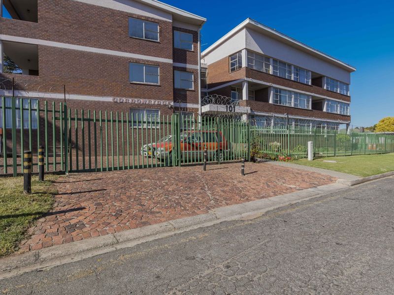 Front exterior view of a multi-story brick apartment building featuring secure perimeter fencing, razor wire, and a paved driveway entrance.
