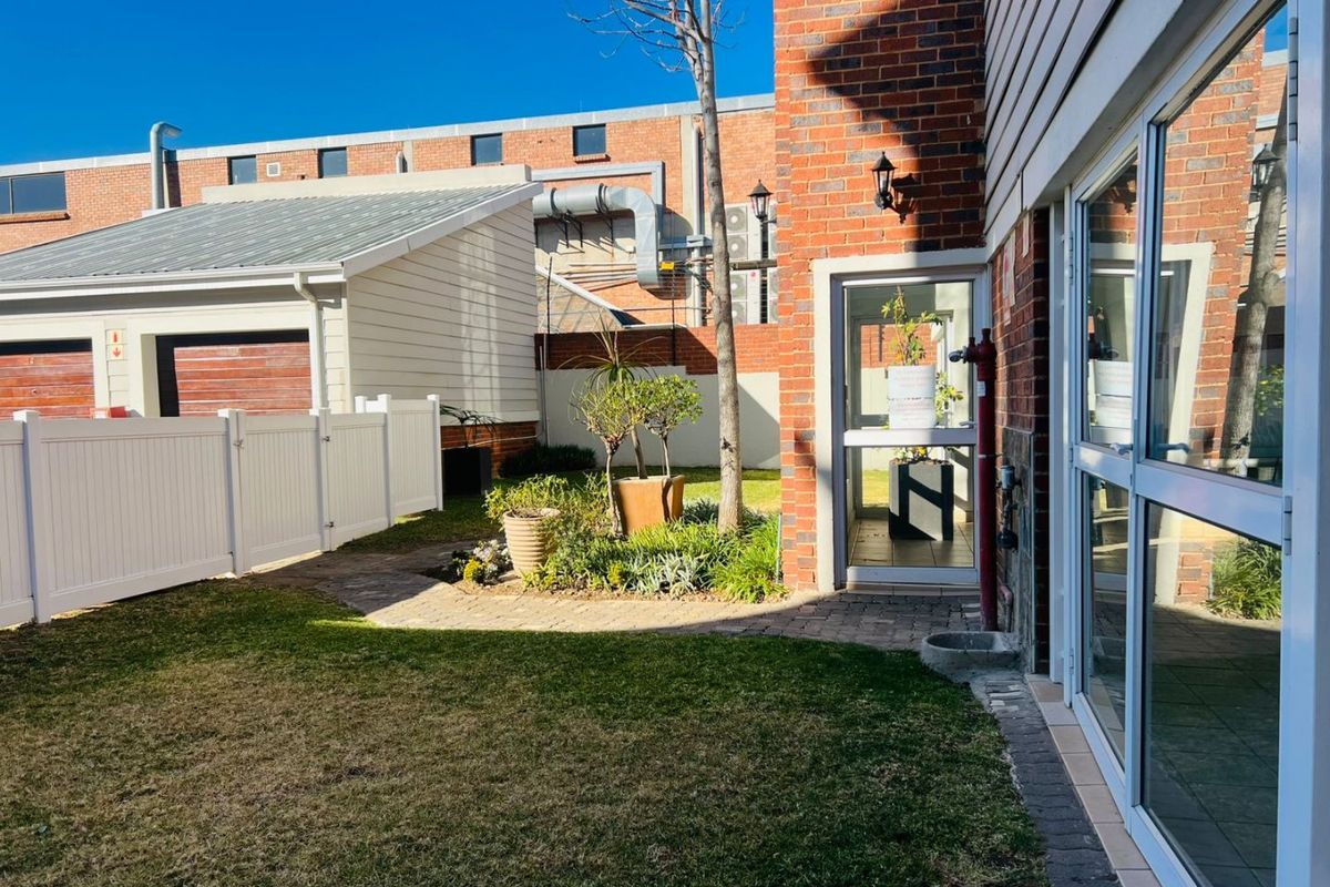 A rear exterior view of the apartment featuring a small garden, paved pathway, brick facade, and sliding glass door access.