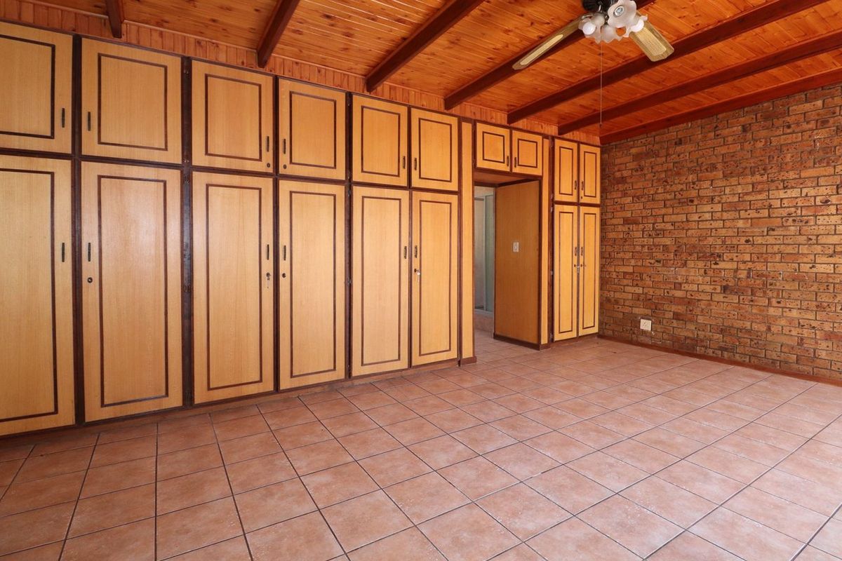 Spacious bedroom featuring extensive built-in cabinetry, exposed timber ceiling beams, a rustic brick feature wall, and durable tiled flooring.