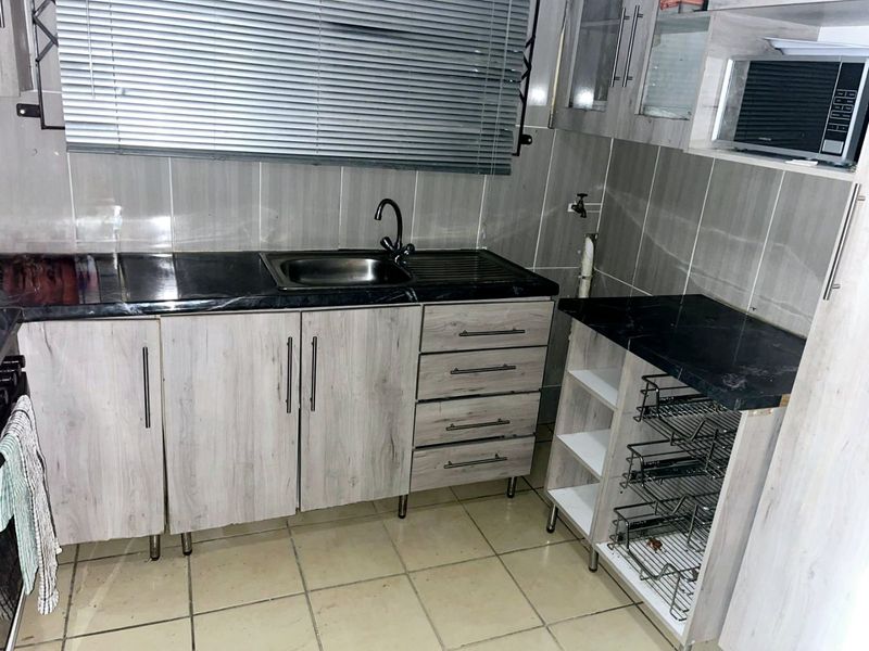 A compact kitchen featuring light wood-grain cabinetry, dark laminate countertops, a stainless steel sink, and integrated storage solutions with tiled wall finishes.