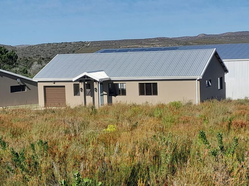 A modern single-story house featuring a corrugated metal roof with integrated solar panels, a garage, and a covered entrance, set in a natural landscape.