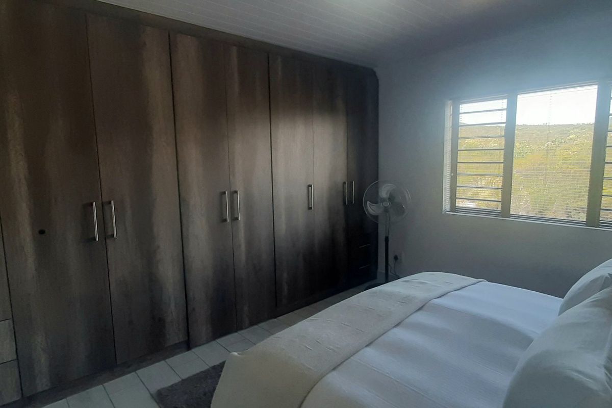 A well-lit bedroom featuring extensive built-in wooden cupboards, tiled flooring, and a window with security bars offering a scenic view.