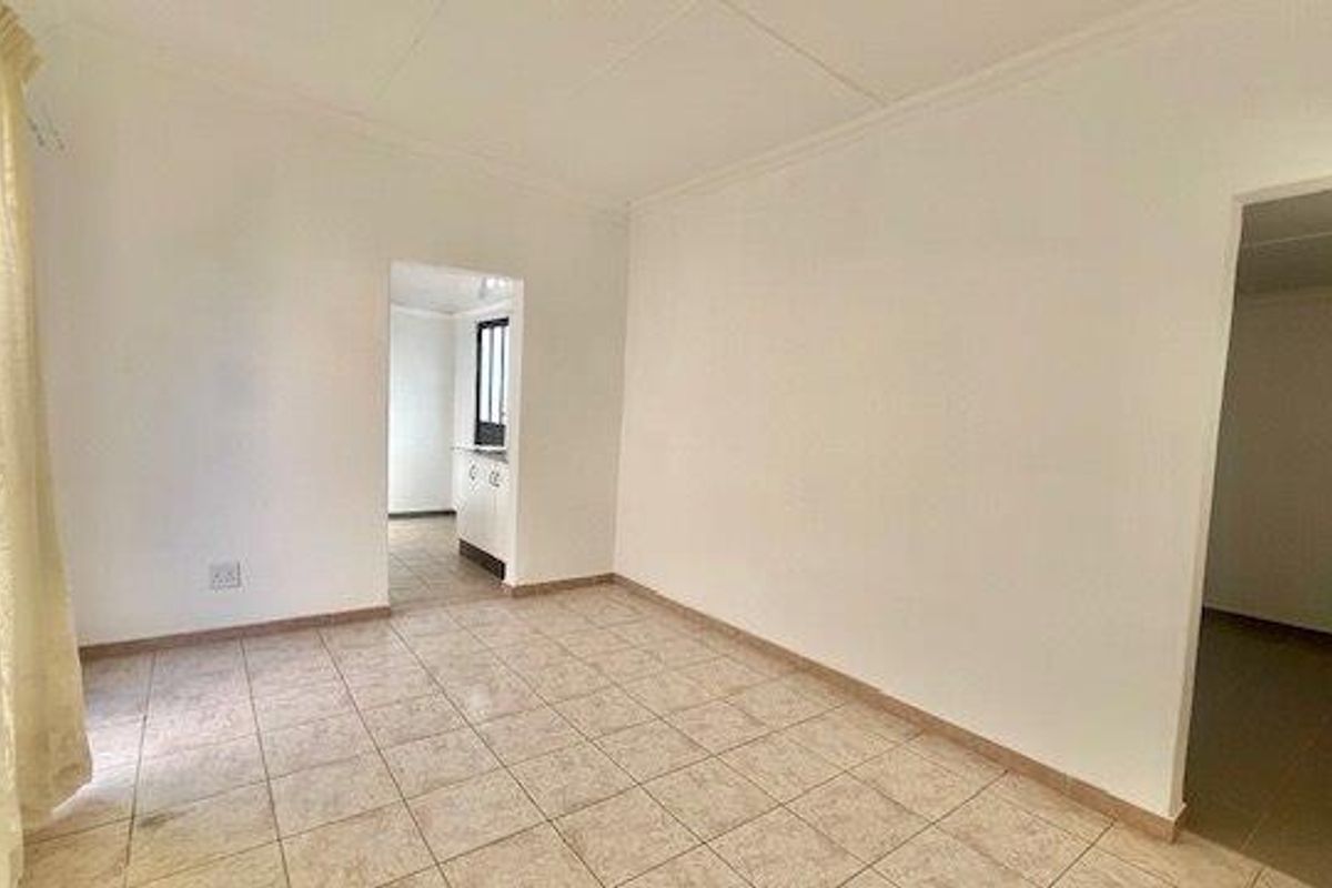 An empty living room featuring tiled flooring and coved ceilings, with an open doorway leading to an adjacent kitchen area.
