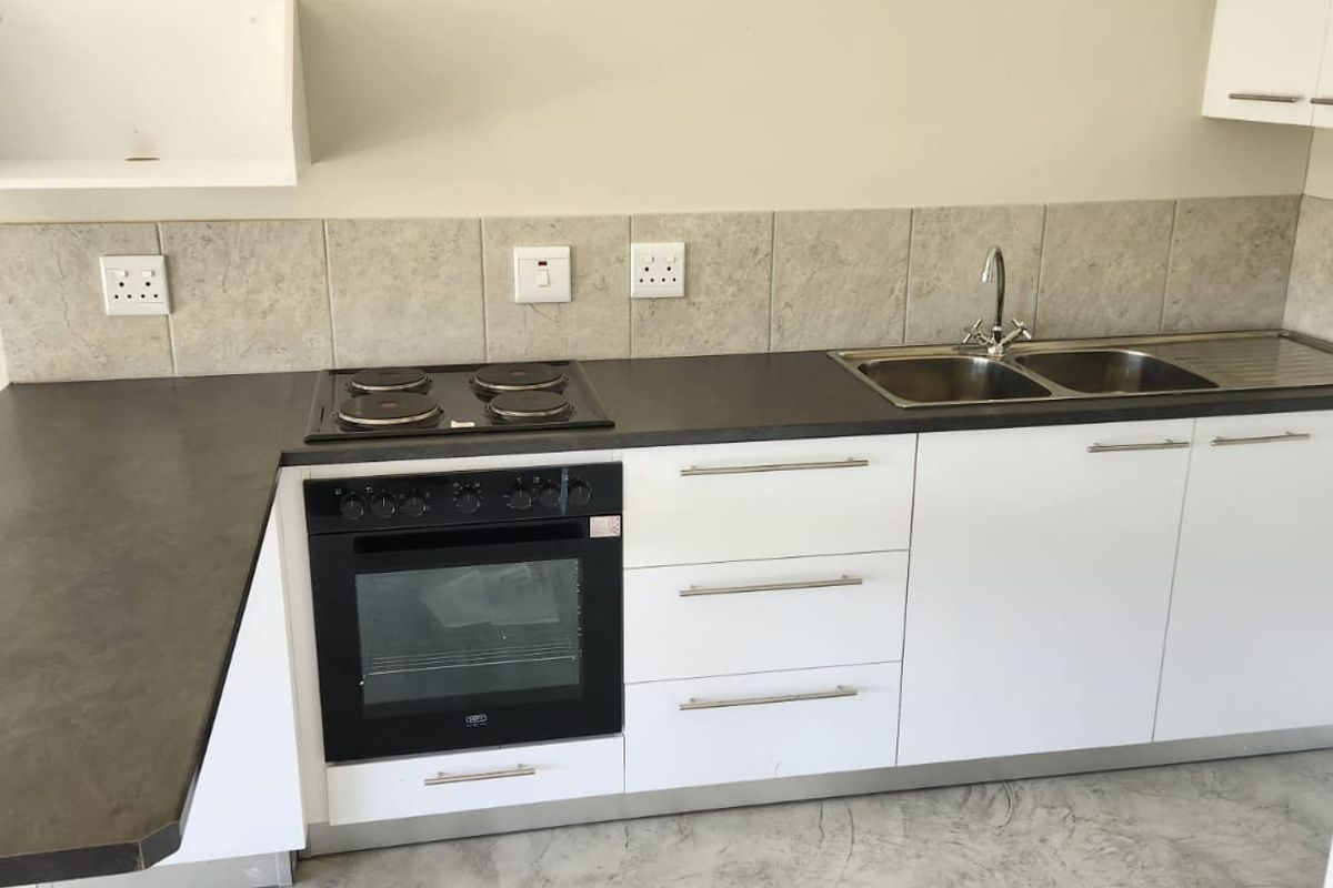 A clean, functional kitchen featuring white cabinetry, dark laminate countertops, a built-in oven, electric hob, and a double sink with tiled backsplash.
