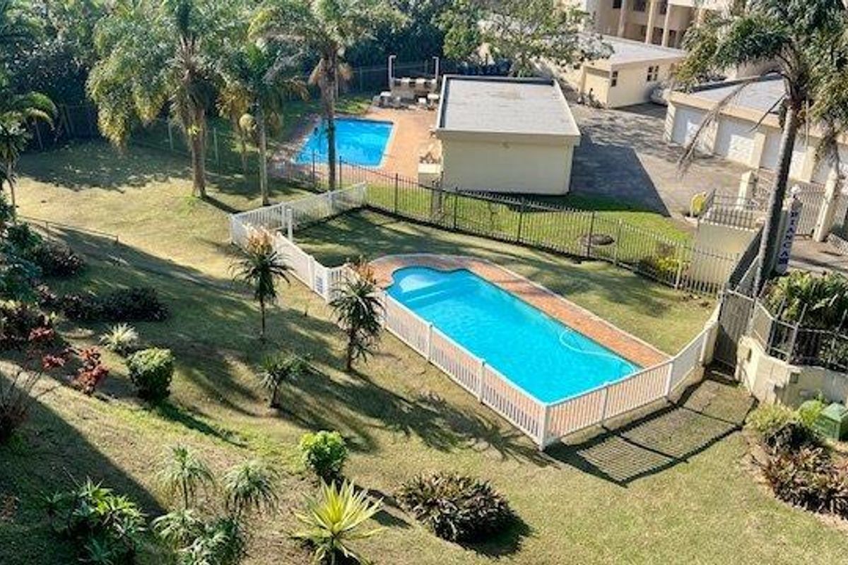 Aerial view of the apartment complex grounds featuring a swimming pool, landscaped garden areas, and a paved driveway with garage access.