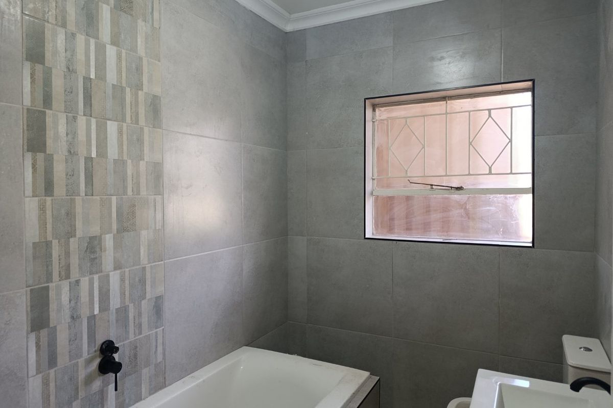 A modern, tiled bathroom featuring a built-in bathtub, pedestal basin, and toilet, finished with contemporary grey wall tiles and decorative accent tiling.