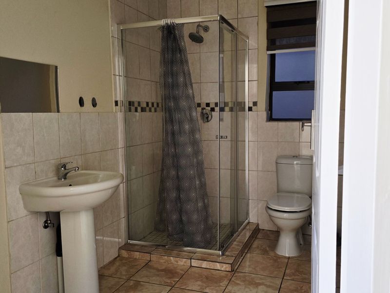 A functional bathroom featuring a pedestal basin, a glass-enclosed shower, and a toilet, finished with neutral wall and floor tiling.