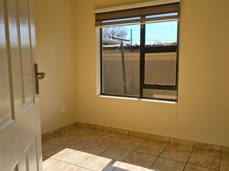 A clean, empty bedroom featuring tiled flooring, neutral walls, and an aluminium-framed window with a roller blind.