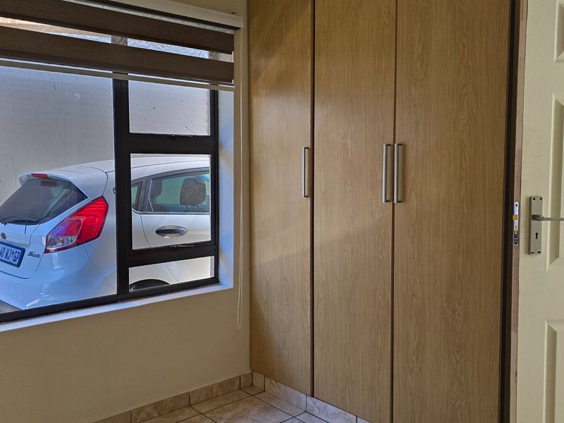 A clean bedroom interior featuring light wood-finish built-in cupboards, tiled flooring, and a large window with a roller blind.