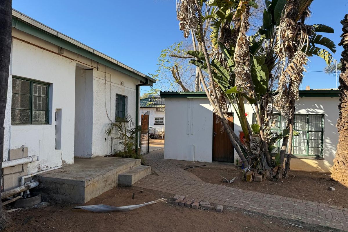 Rear exterior view of the property featuring a paved walkway, concrete porch, and established garden area with mature trees.