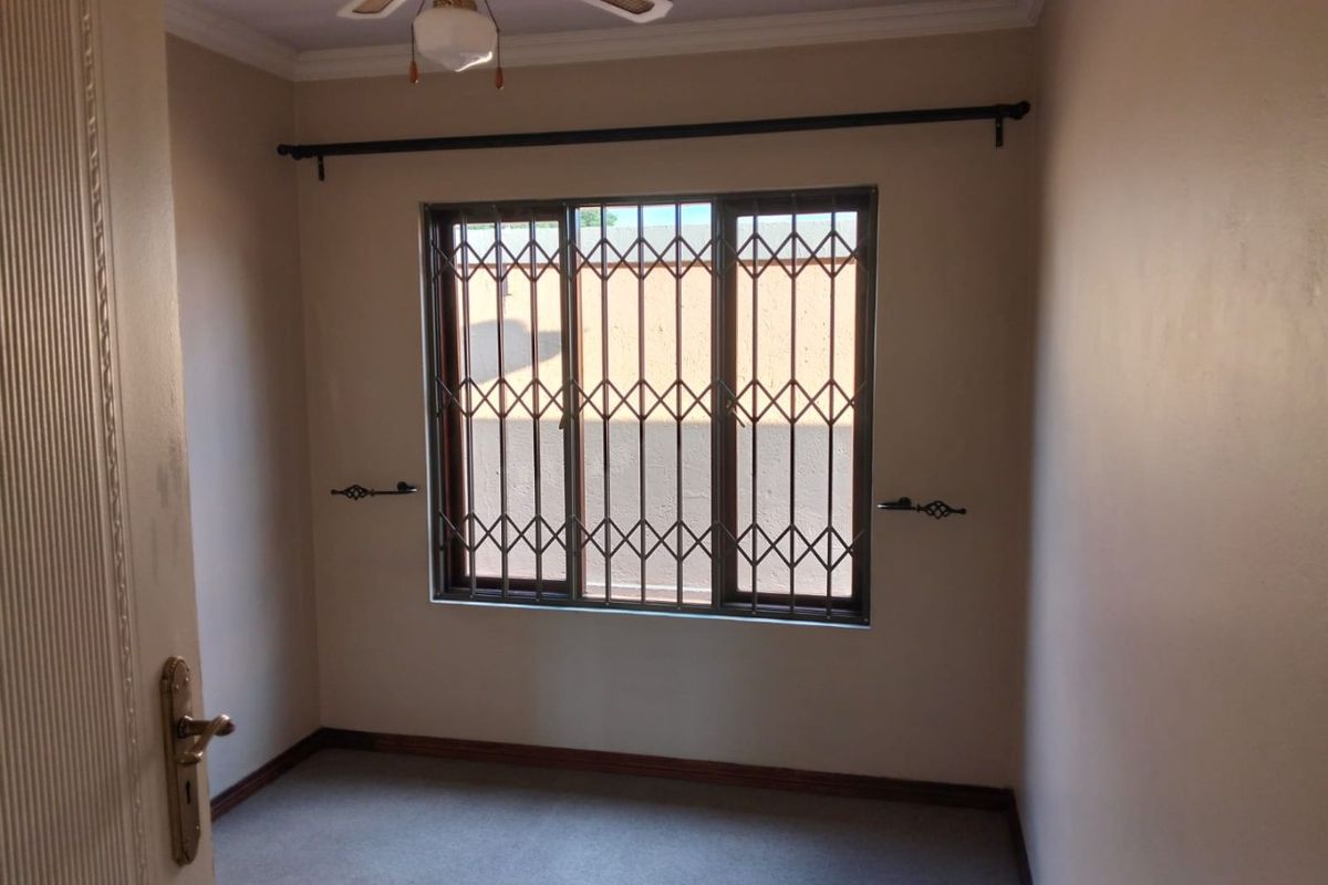 An empty bedroom featuring neutral walls, carpeted flooring, a ceiling fan, and a window fitted with a security gate.