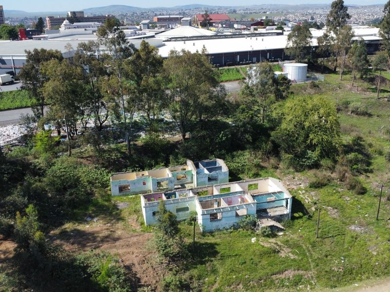 Aerial view of a vacant residential plot featuring a derelict, roofless structure surrounded by overgrown vegetation and industrial surroundings.