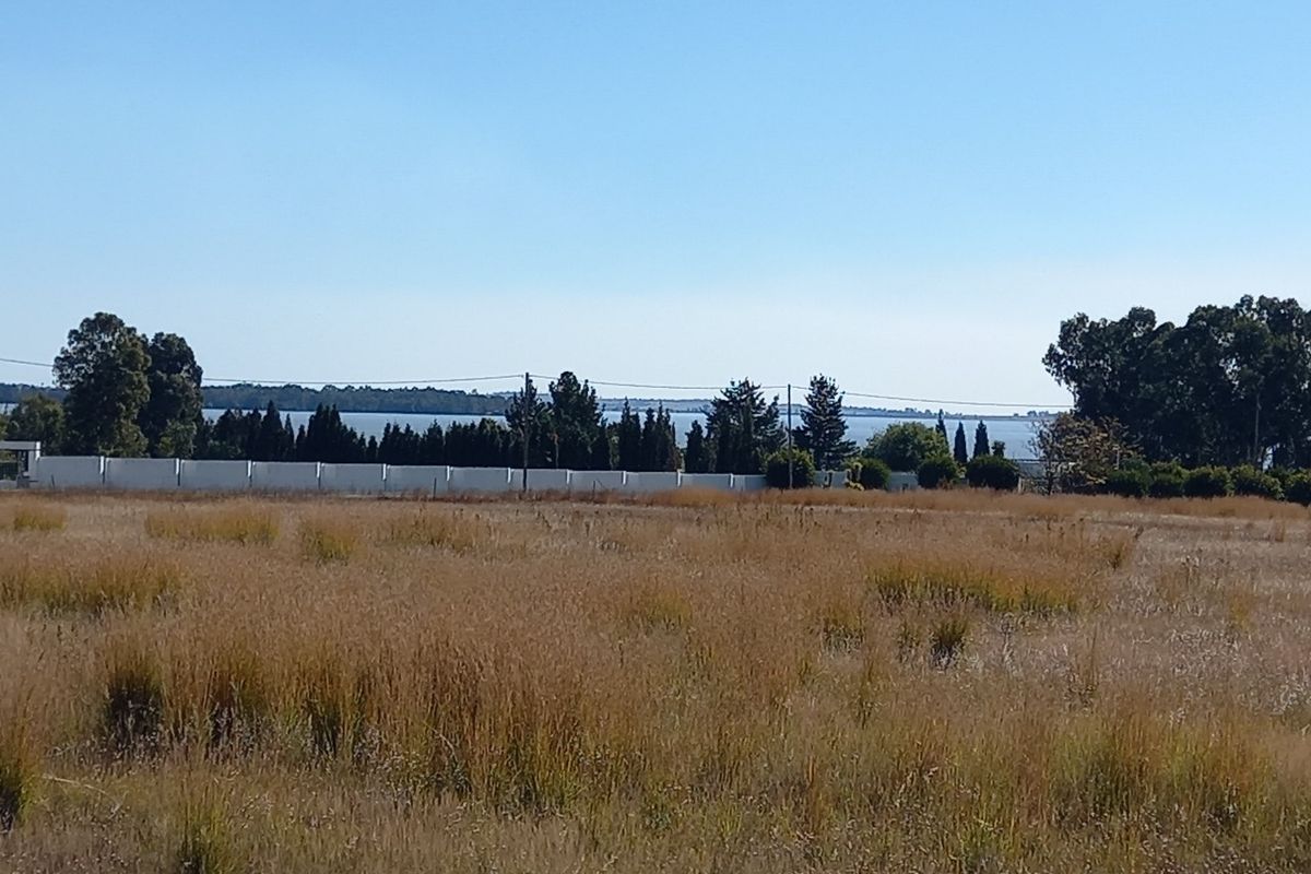 A view of a vacant residential plot featuring dry grass, a perimeter wall, and distant water views.