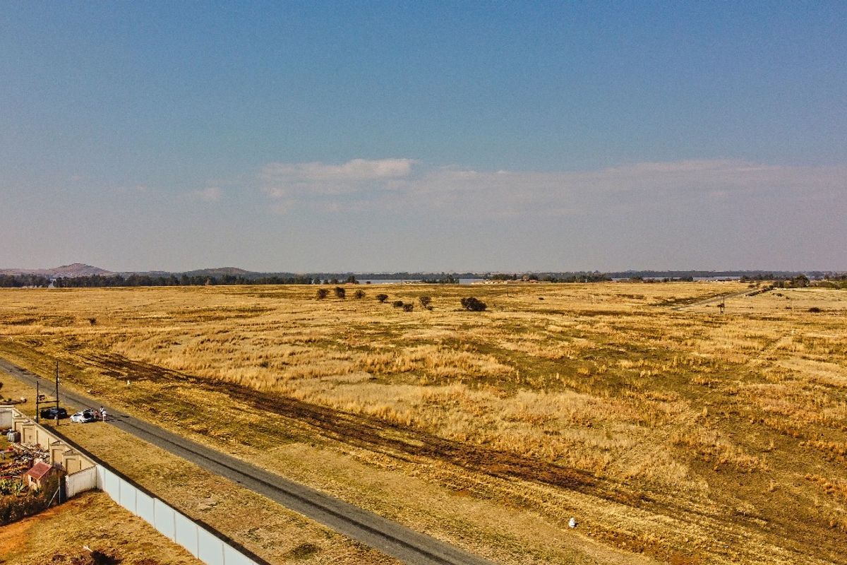 An aerial view of a large, flat, vacant residential plot featuring dry grassland, perimeter fencing, and adjacent road access.