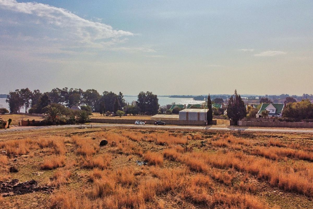An elevated aerial view of a vacant residential plot featuring dry grass, perimeter fencing, and distant water views.