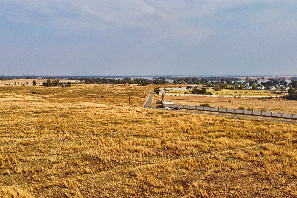 An aerial view of a large, undeveloped residential land parcel featuring dry grass, a boundary wall, and access to a nearby paved road.