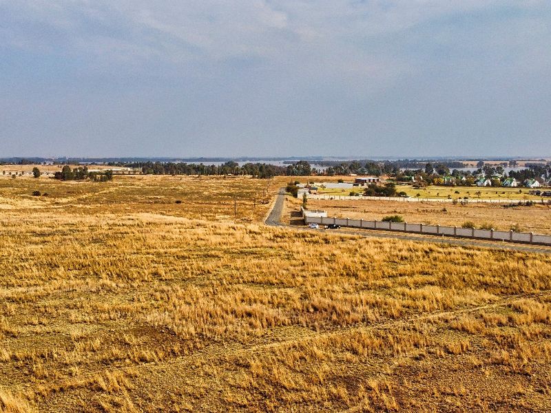 An aerial view of a large, undeveloped residential land parcel featuring dry grass, adjacent to a paved road and existing perimeter walling.