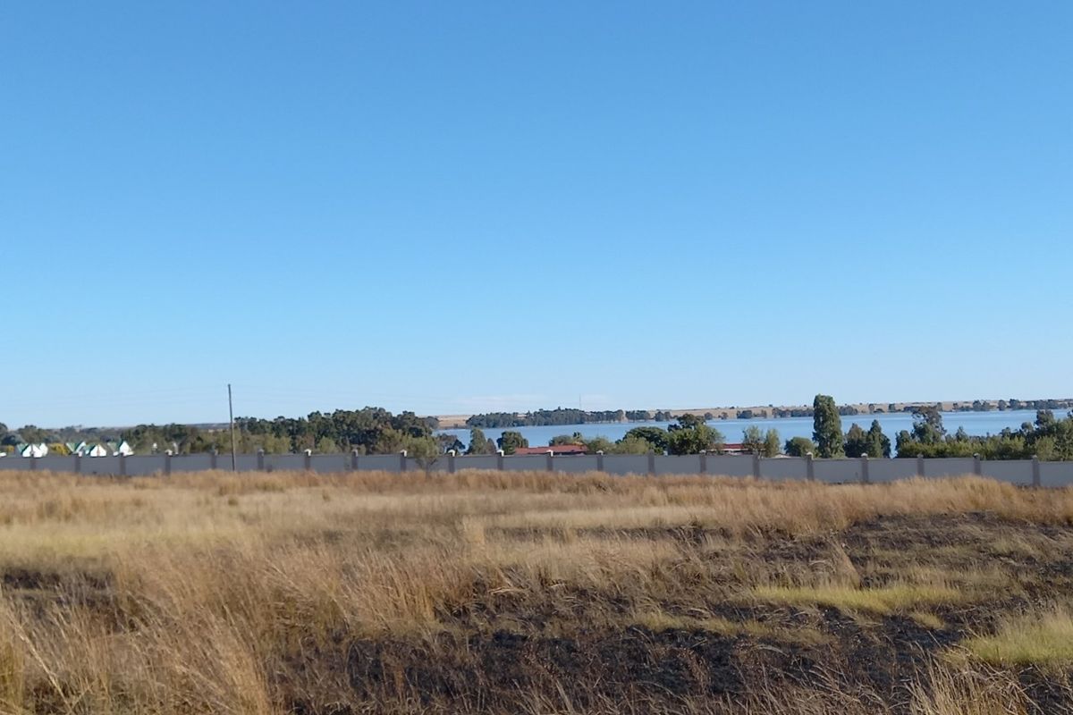 A view of the vacant residential land featuring dry grass and a perimeter wall, with a scenic lake view in the background.
