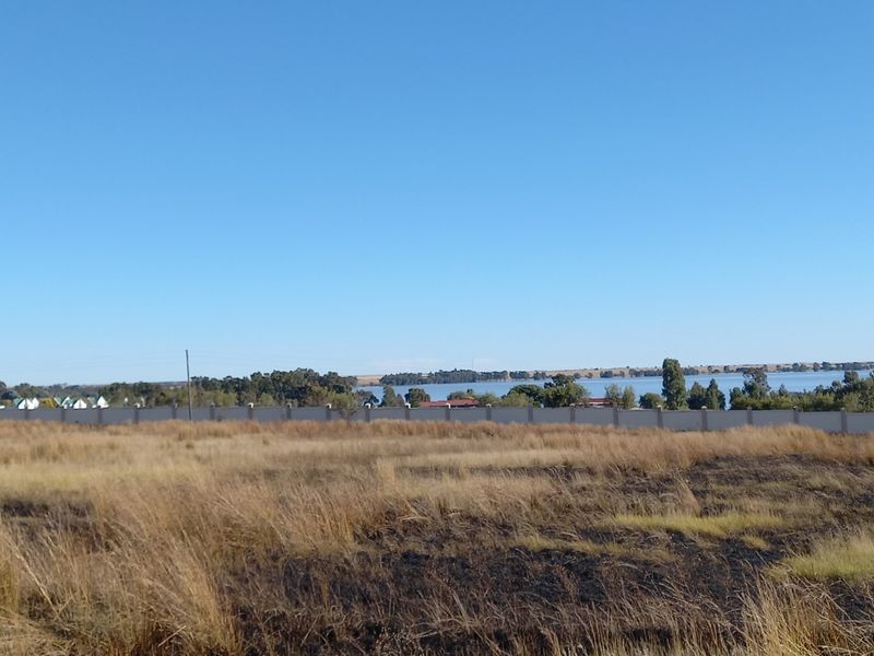 A view of the vacant residential land featuring dry grass and a perimeter wall, with a scenic lake view in the background.