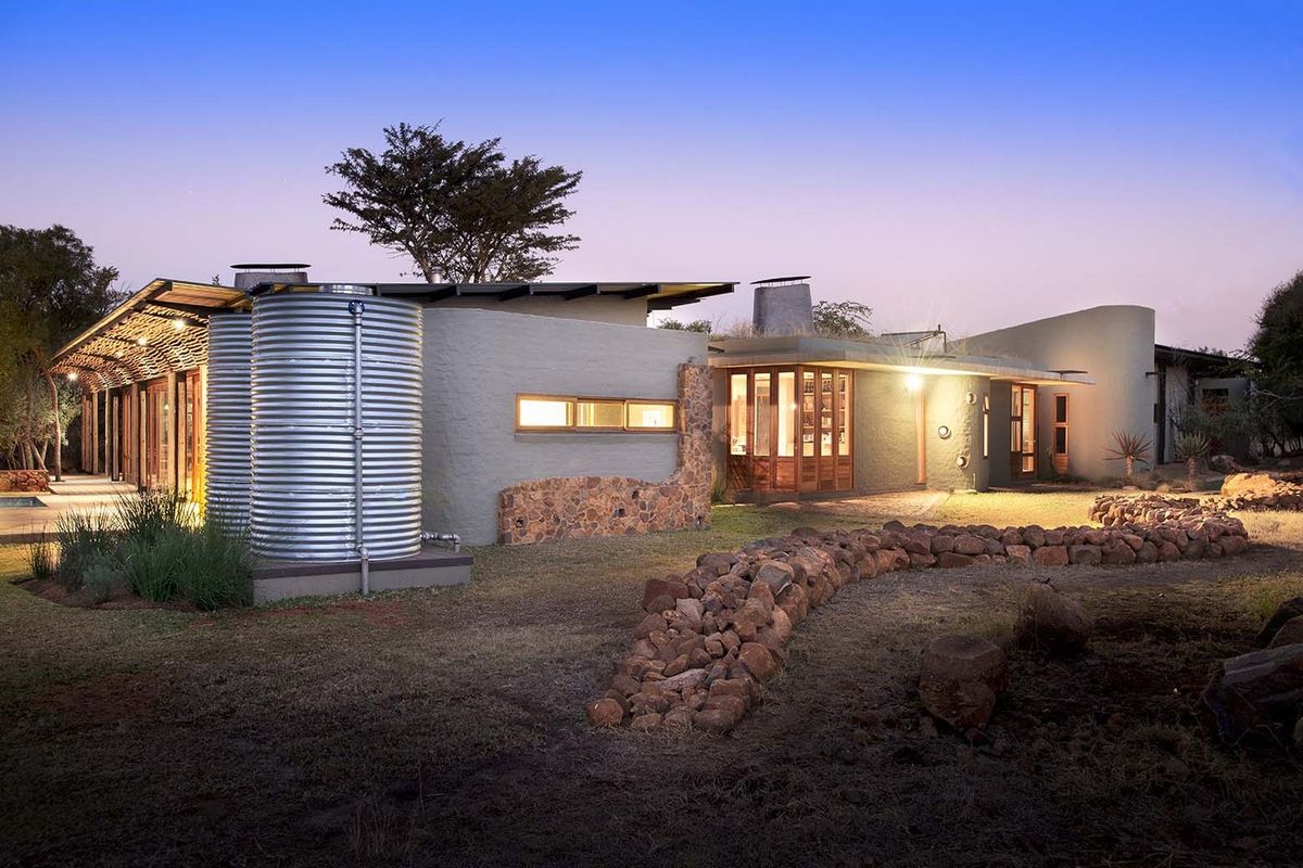 A modern architectural residence featuring stone accents, corrugated metal water tanks, and expansive glass windows set within a natural landscape.