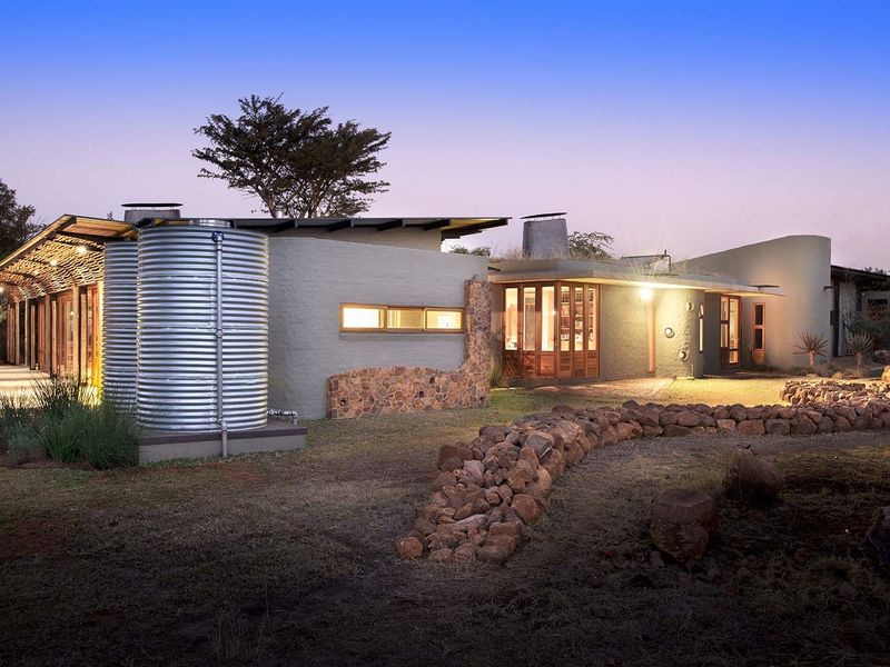 A modern architectural residence featuring stone accents, corrugated metal water tanks, and expansive glass windows set within a natural landscape.