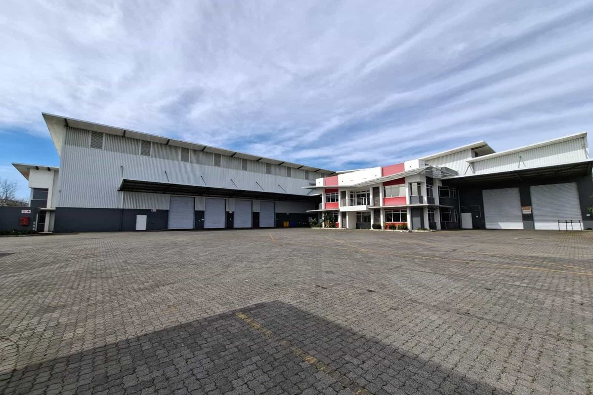 Wide-angle view of an industrial warehouse facility featuring multiple roller shutter doors, a large paved yard, and a modern office building section.