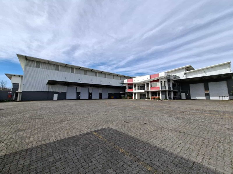 Wide-angle view of an industrial warehouse facility featuring multiple roller shutter doors, a large paved yard, and a modern office building section.