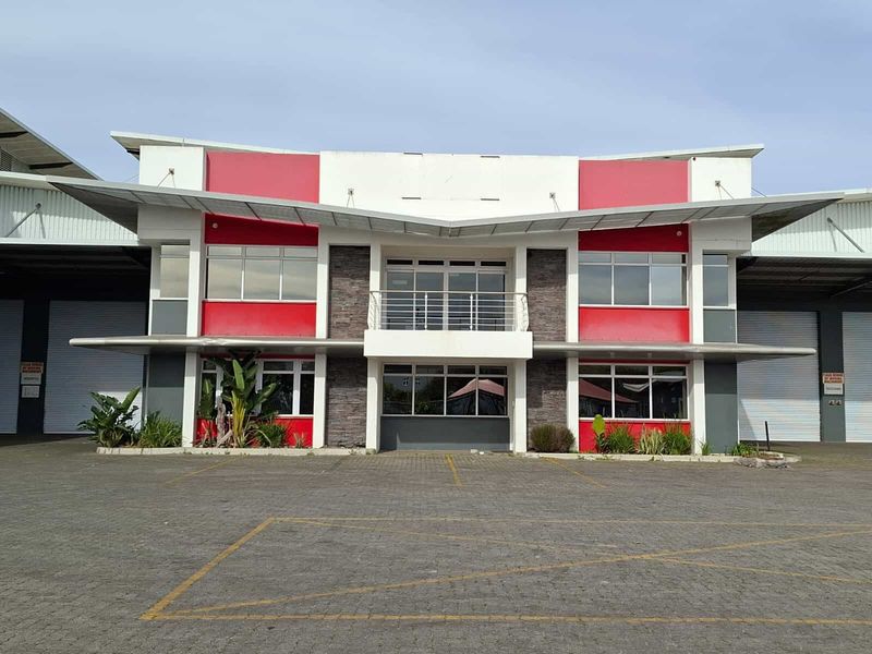 Front elevation of a modern industrial facility featuring a central office block with balcony, flanked by large warehouse sections with roller shutter doors.