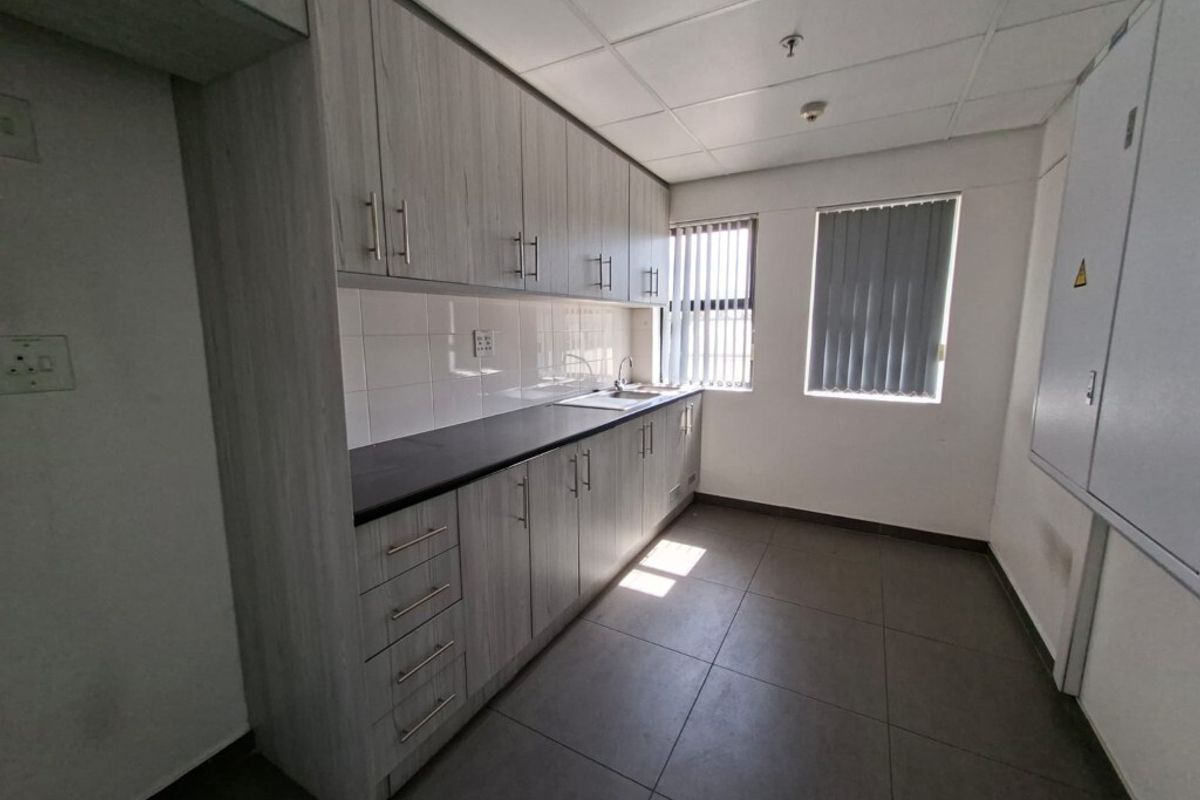 A functional kitchenette featuring light wood-grain cabinetry, dark countertops, a stainless steel sink, and tiled flooring in an industrial office setting.