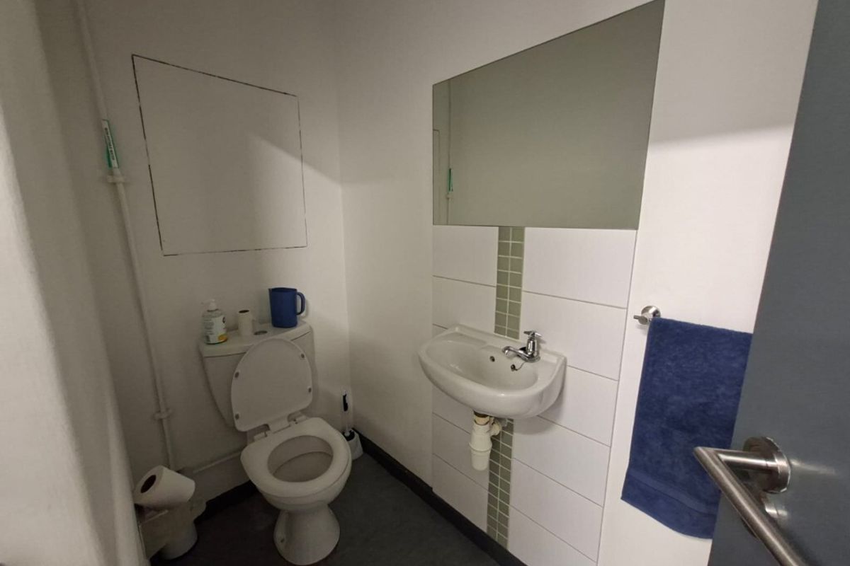 A functional industrial-style restroom featuring a wall-mounted basin, a standard toilet, and a large wall mirror with tiled splashback.