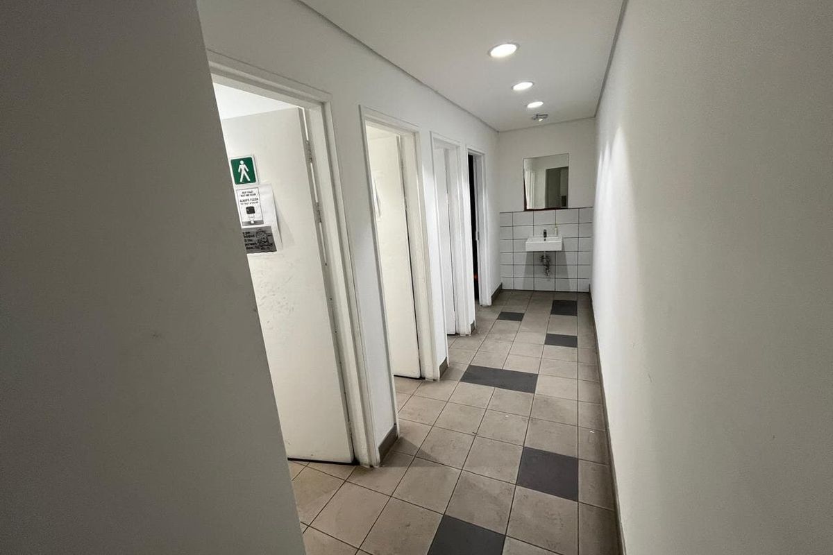 A functional industrial-style bathroom corridor featuring tiled flooring, recessed lighting, and multiple cubicle access points with a wall-mounted basin.
