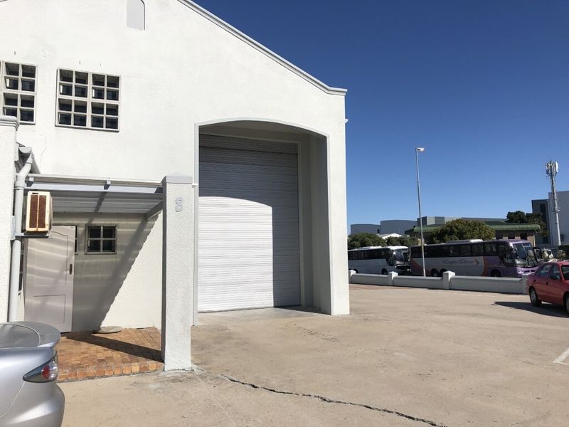 Industrial property exterior featuring a large roller shutter door, concrete driveway, and paved entrance area under a clear blue sky.