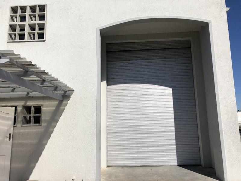 Industrial property exterior featuring a large roller shutter door, concrete facade, and a decorative breeze block window with an attached pergola.