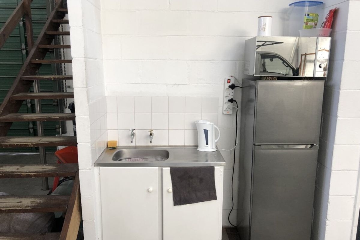 A basic industrial kitchenette featuring a stainless steel sink, tiled splashback, and built-in cabinetry, situated adjacent to a timber staircase.