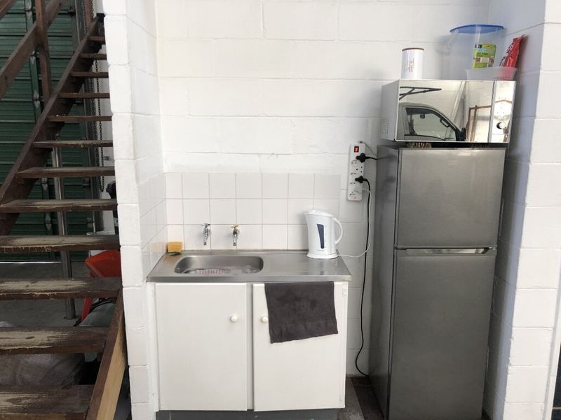 A basic industrial kitchenette featuring a stainless steel sink, tiled splashback, and built-in cabinetry, situated adjacent to a timber staircase.