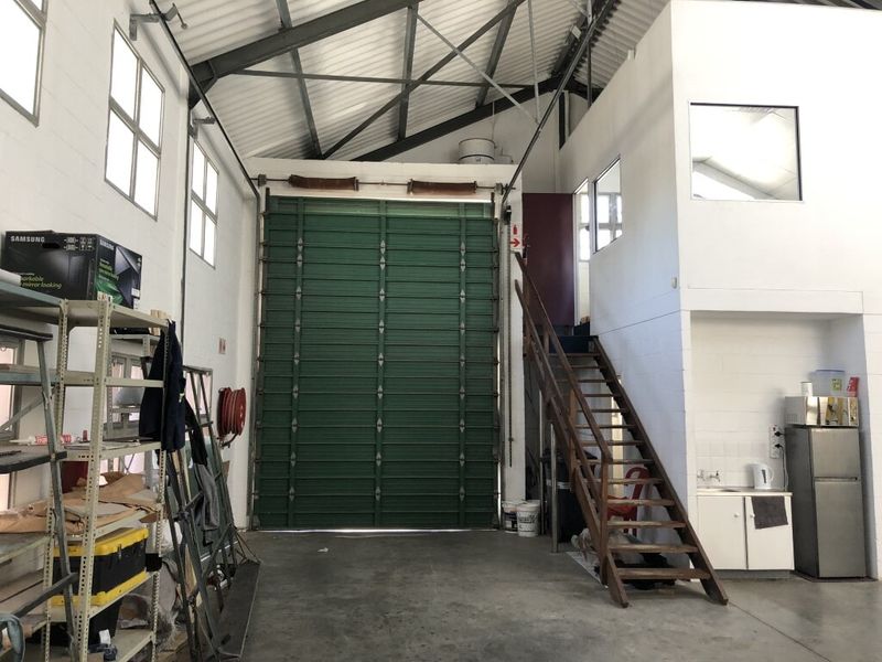 Industrial warehouse space featuring a large roller shutter door, exposed steel roof trusses, a mezzanine level with timber staircase, and a functional kitchenette area.