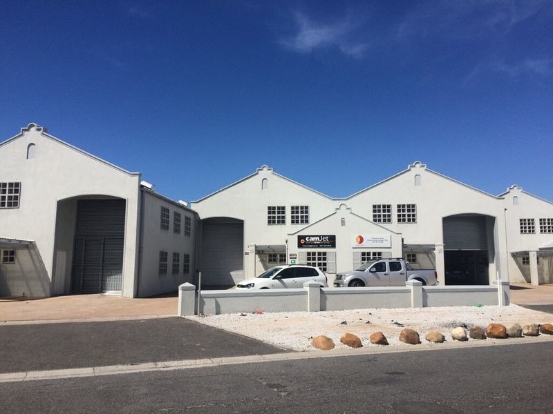 Front elevation of an industrial complex featuring multiple units with roller shutter doors, gabled roof architecture, and a paved driveway.