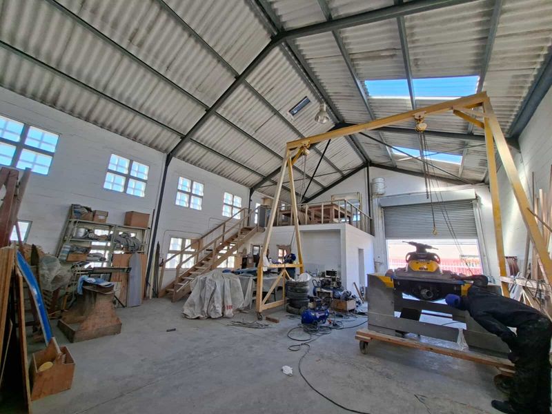 Spacious industrial workshop featuring high ceilings with exposed steel trusses, a mezzanine level, gantry crane, and large roller shutter door access.