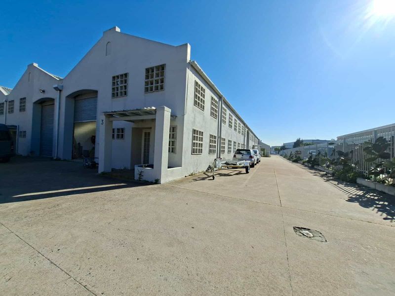 Front elevation of an industrial warehouse unit featuring a roller shutter door, concrete driveway, and secure perimeter fencing.