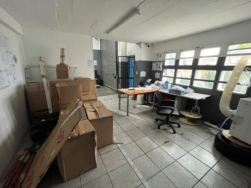Functional industrial office space featuring tiled flooring, large windows for natural light, and direct access to the warehouse area via a roller shutter door.