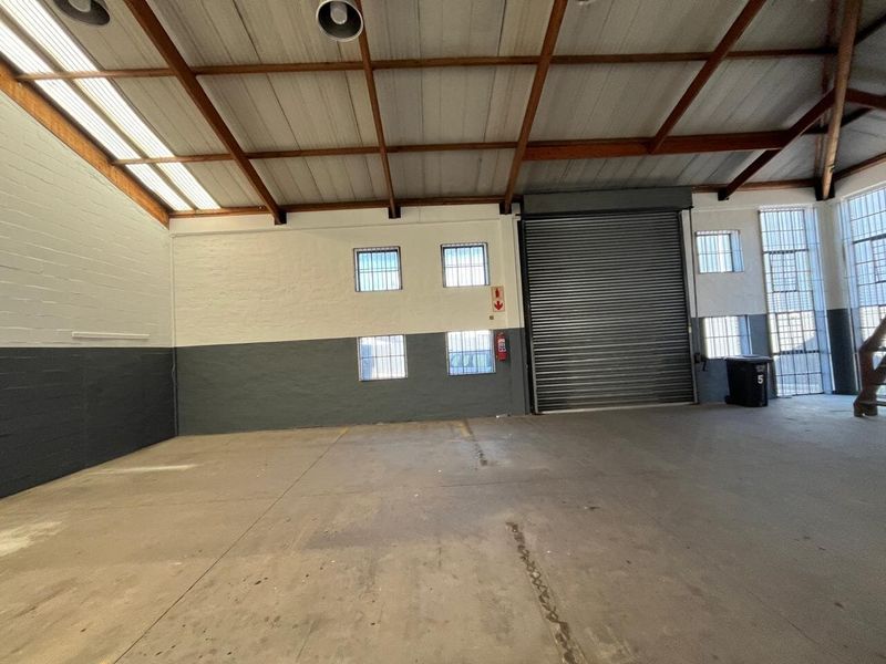Spacious industrial utility area featuring high ceilings with exposed timber trusses, concrete flooring, a large roller shutter door, and secure window grilles.