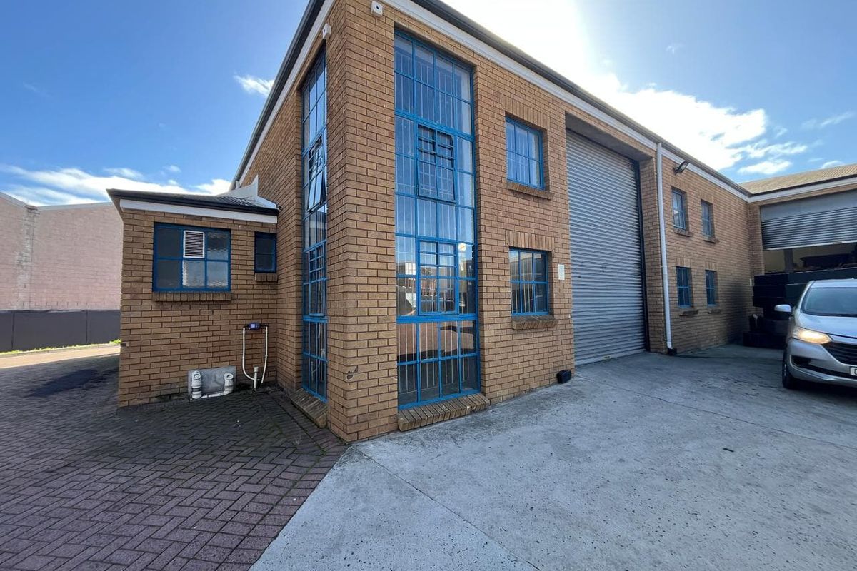 Industrial property exterior featuring a brick facade, large roller shutter door, and blue-framed steel windows with a concrete driveway and paved entrance area.