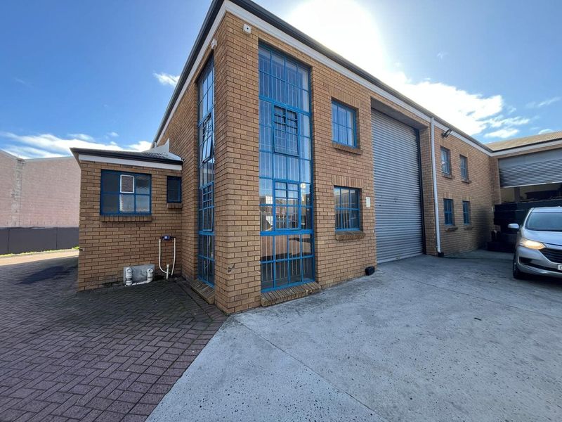 Industrial property exterior featuring a brick facade, large roller shutter door, and blue-framed steel windows with a concrete driveway and paved entrance area.