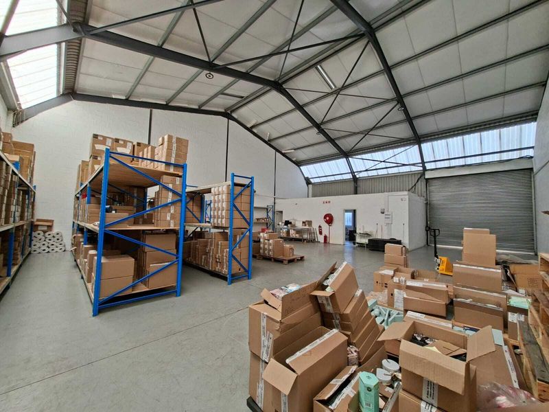 Spacious industrial warehouse unit featuring high ceilings with exposed steel trusses, concrete flooring, and a large roller shutter door for easy access.